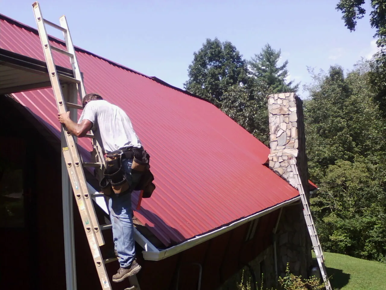 Roofer on a ladder beside a red metal roof and stone chimney — Soffit & Fascia Repair by Roofing Brothers in Thorofare